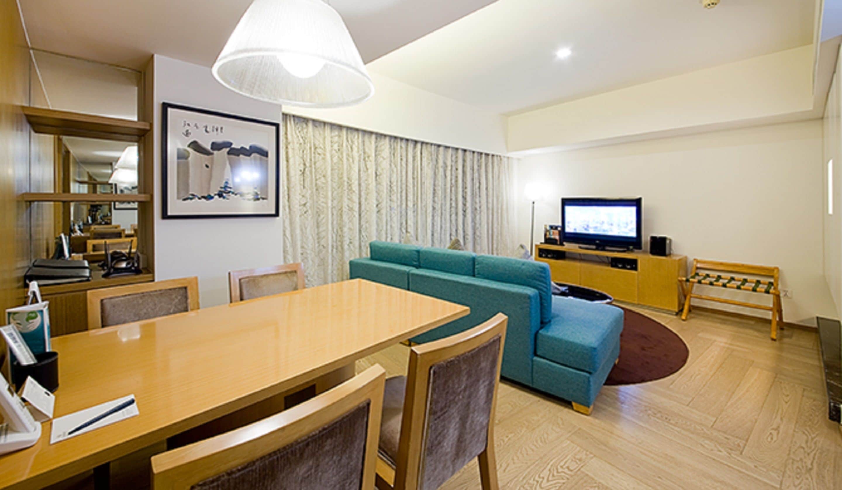 A modern living room with a wooden dining table, teal sofa, and a television on a wooden stand.