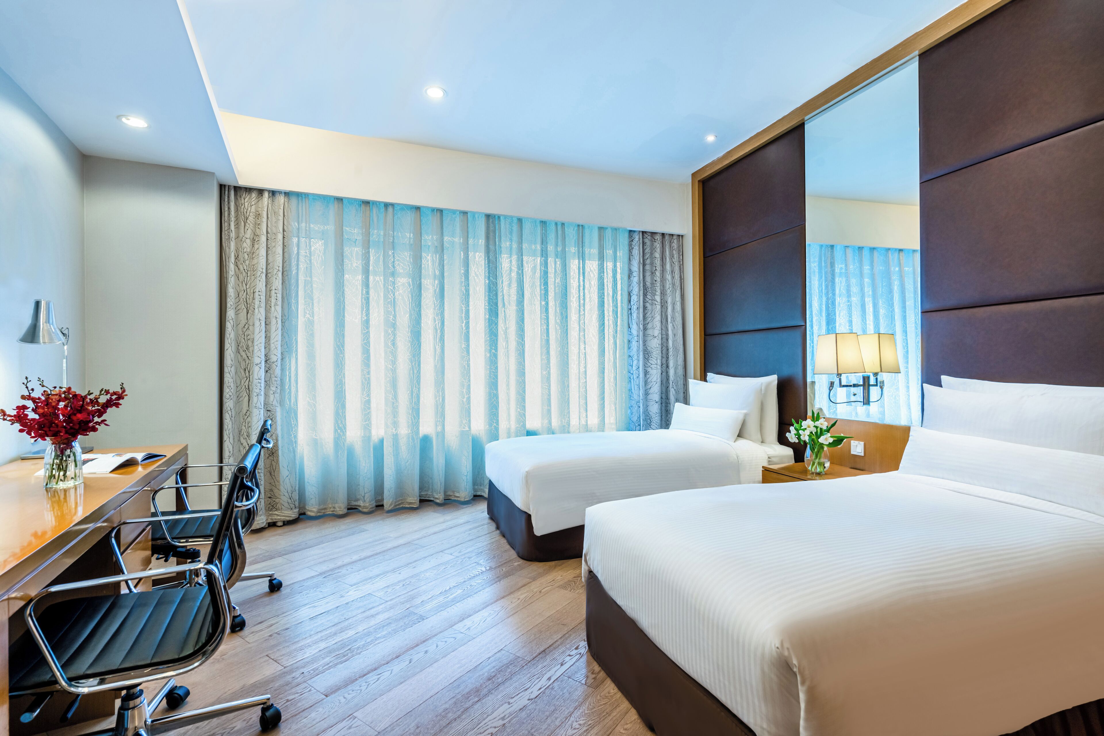A hotel room with two beds, a desk with a chair, a lamp, and a vase with flowers.
