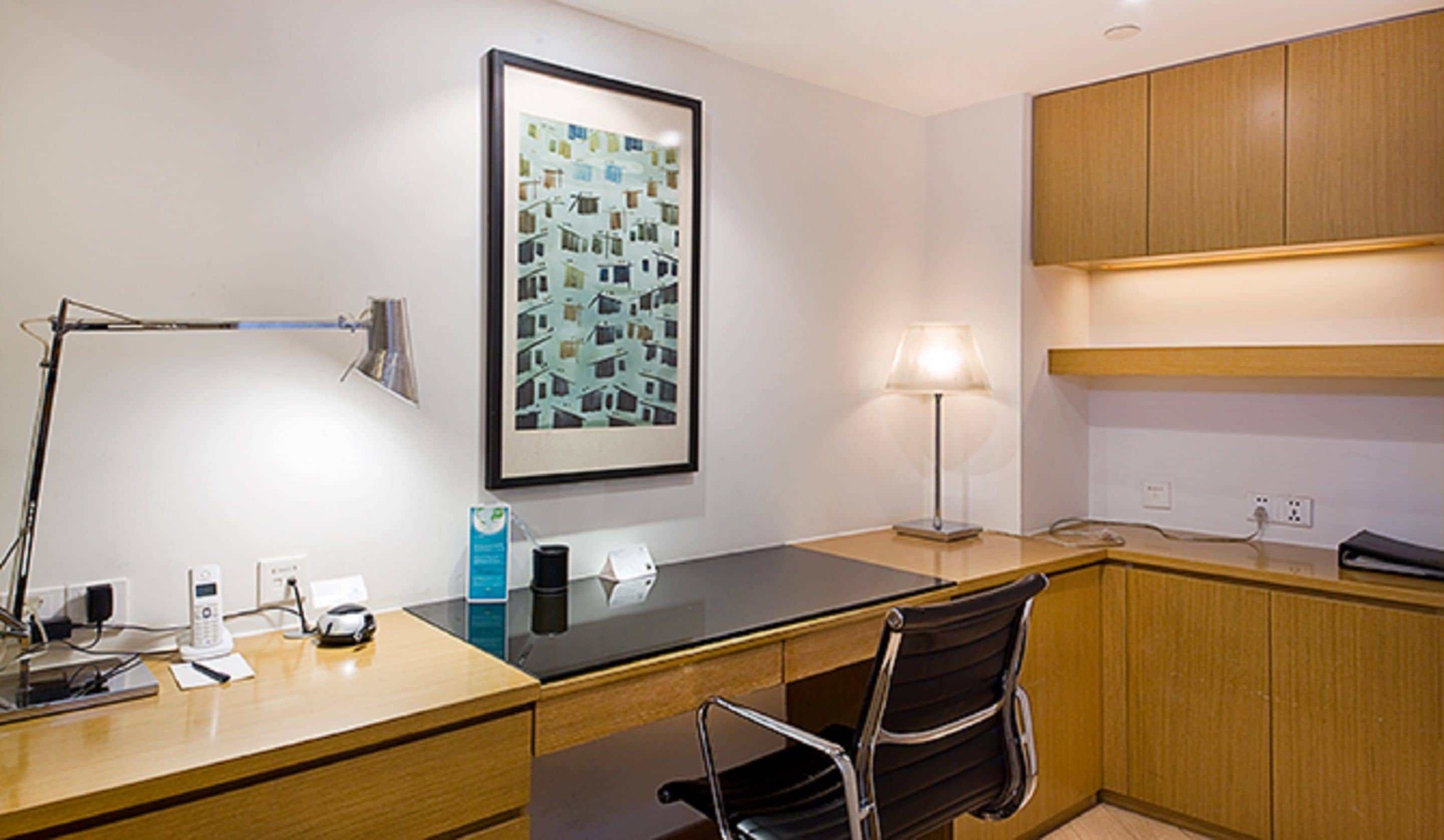 A modern hotel room with a wooden desk, a black chair, a framed picture on the wall, a lamp, and a phone.