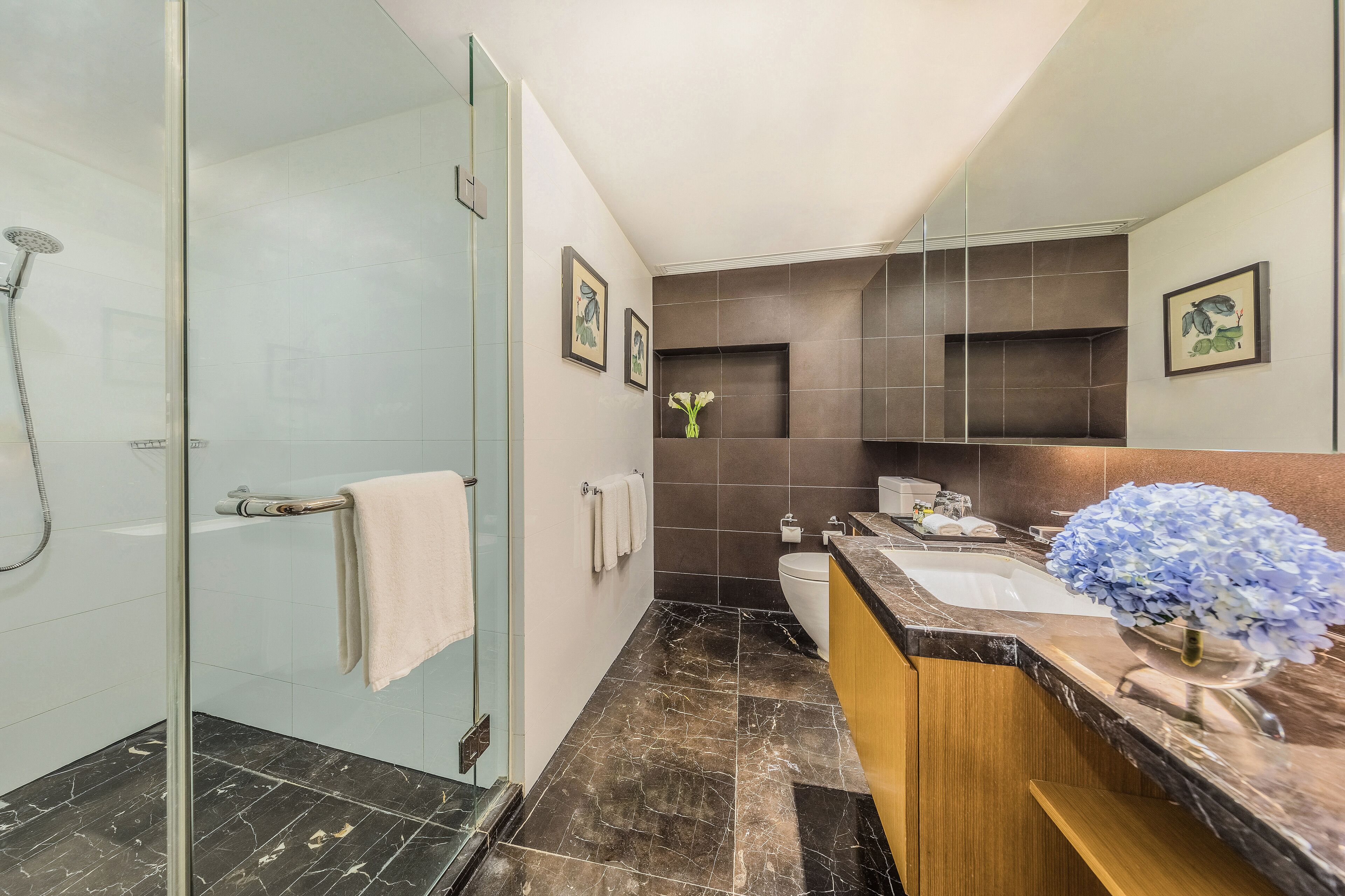 Un baño moderno con cabina de ducha de vidrio, suelo de mármol, tocador de madera con lavamanos y un espejo grande.