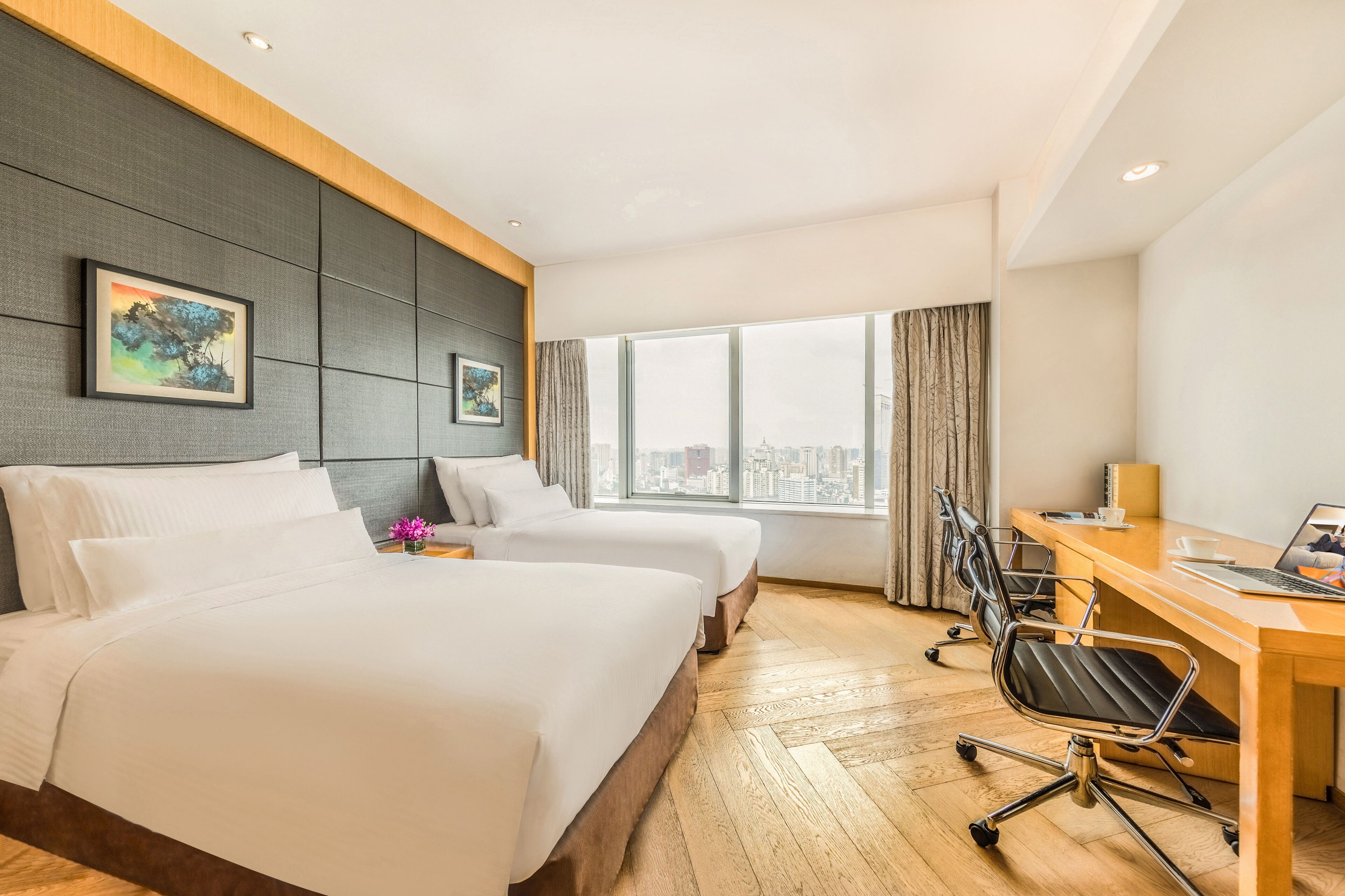 A hotel room with two beds, a desk, and a large window offering a city view.