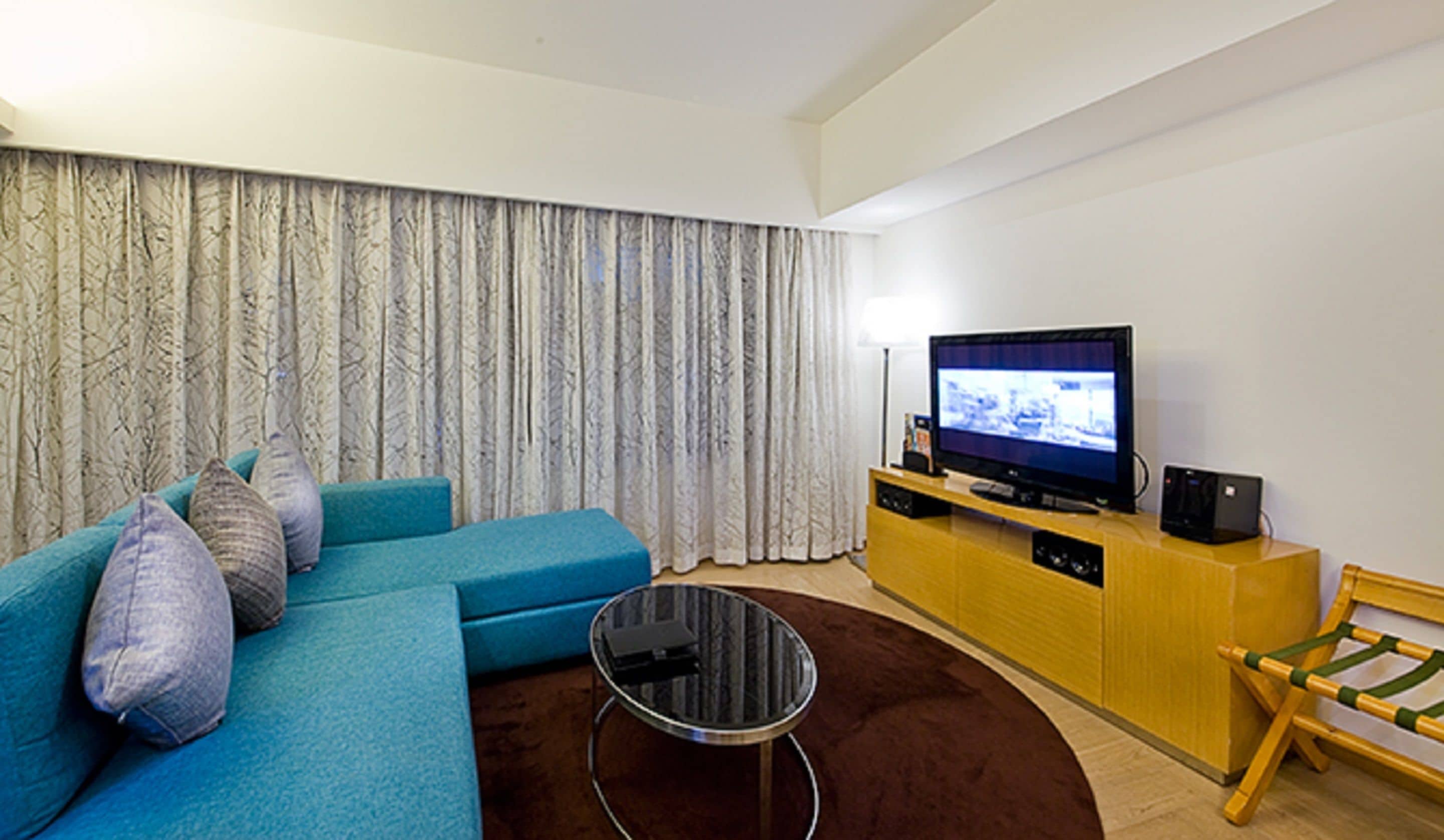 Een moderne woonkamer met een blauwe bank, een klein rond bijzettafeltje, een televisie op een houten meubel en een groot raam met gordijnen.