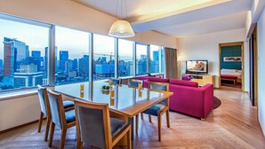 Una habitación de hotel moderna con zona de comedor, sofá rosa, televisor y ventana grande con vistas a la ciudad.
