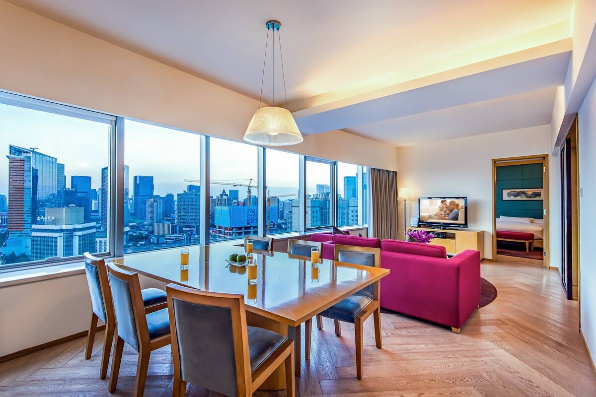 Ett modernt hotellrum med en matplats, en rosa soffa, en TV och ett stort fönster med utsikt över staden.