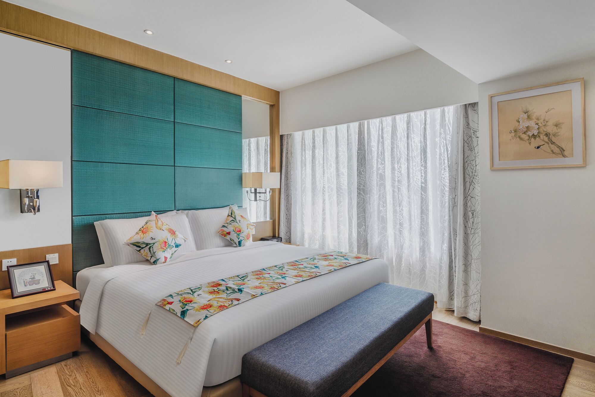 A hotel room with a large bed, a bench, a nightstand with a lamp, and a framed picture on the wall.