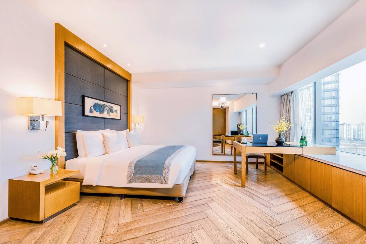 Ett modernt hotellrum med en stor säng, ett skrivbord med en bärbar dator, utsikt över staden genom stora fönster och ett trägolv.