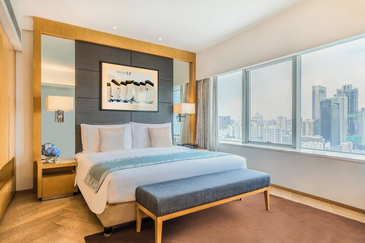 Ett modernt hotellrum med en stor säng, en bänk, utsikt över staden och ett inramat konstverk på väggen.