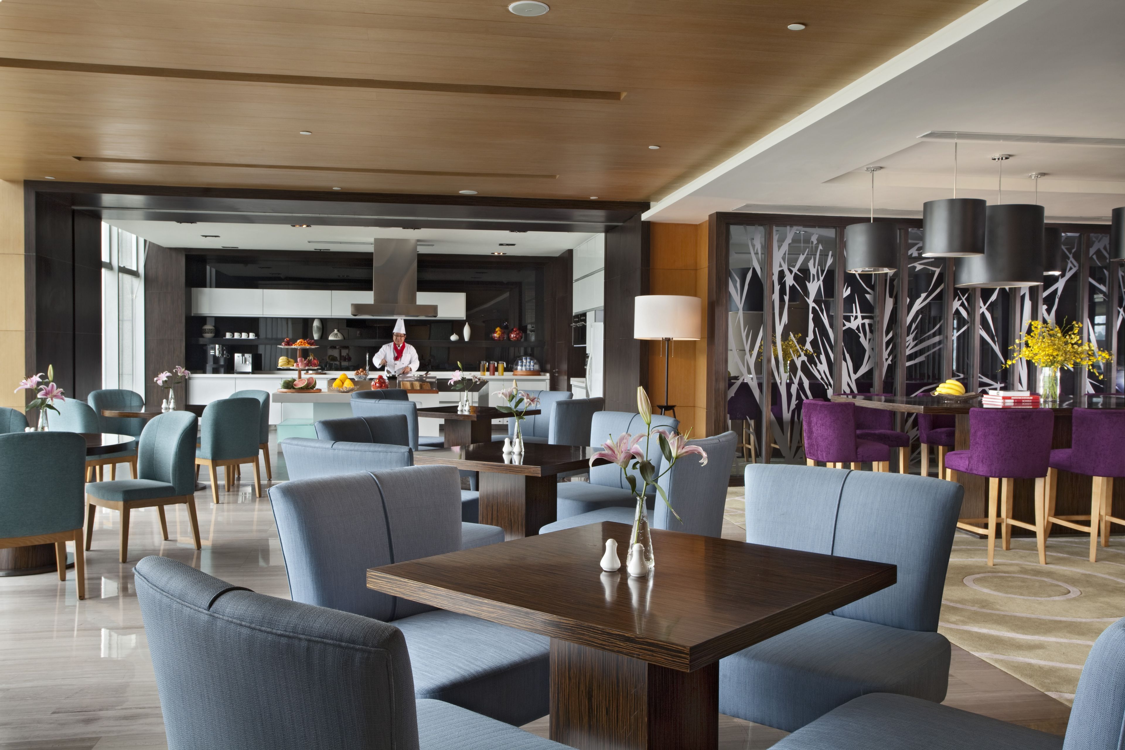 A modern hotel lobby with a central seating area, a bar, and a kitchen in the background.