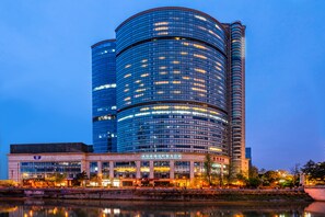 Un hotel moderno con fachada de vidrio curva, una señal con el nombre a la vista y un reflejo en el agua.