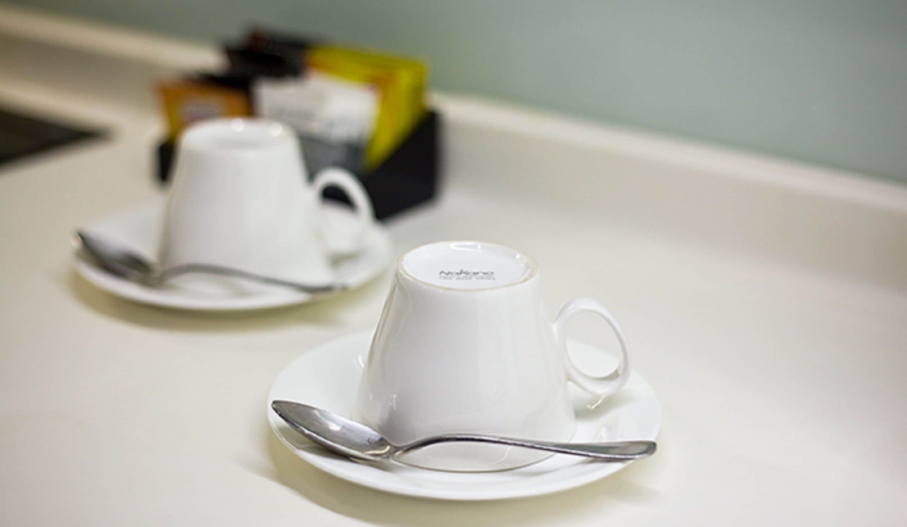 Two white cups on saucers with spoons, placed on a white table.