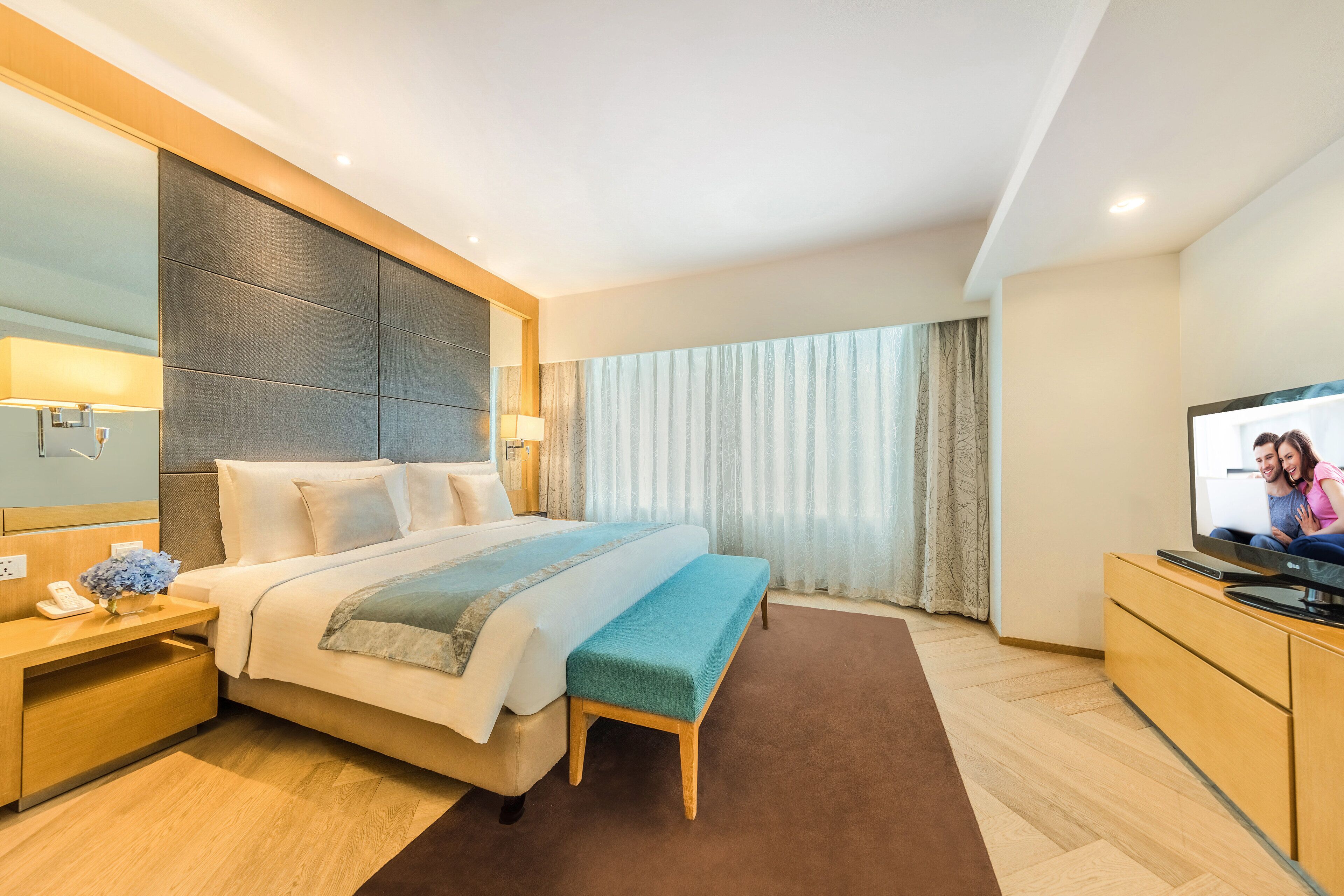 Una habitación de hotel moderna con una cama grande, una mesita de noche con lámpara, un televisor sobre un mueble de madera y una ventana con cortinas.