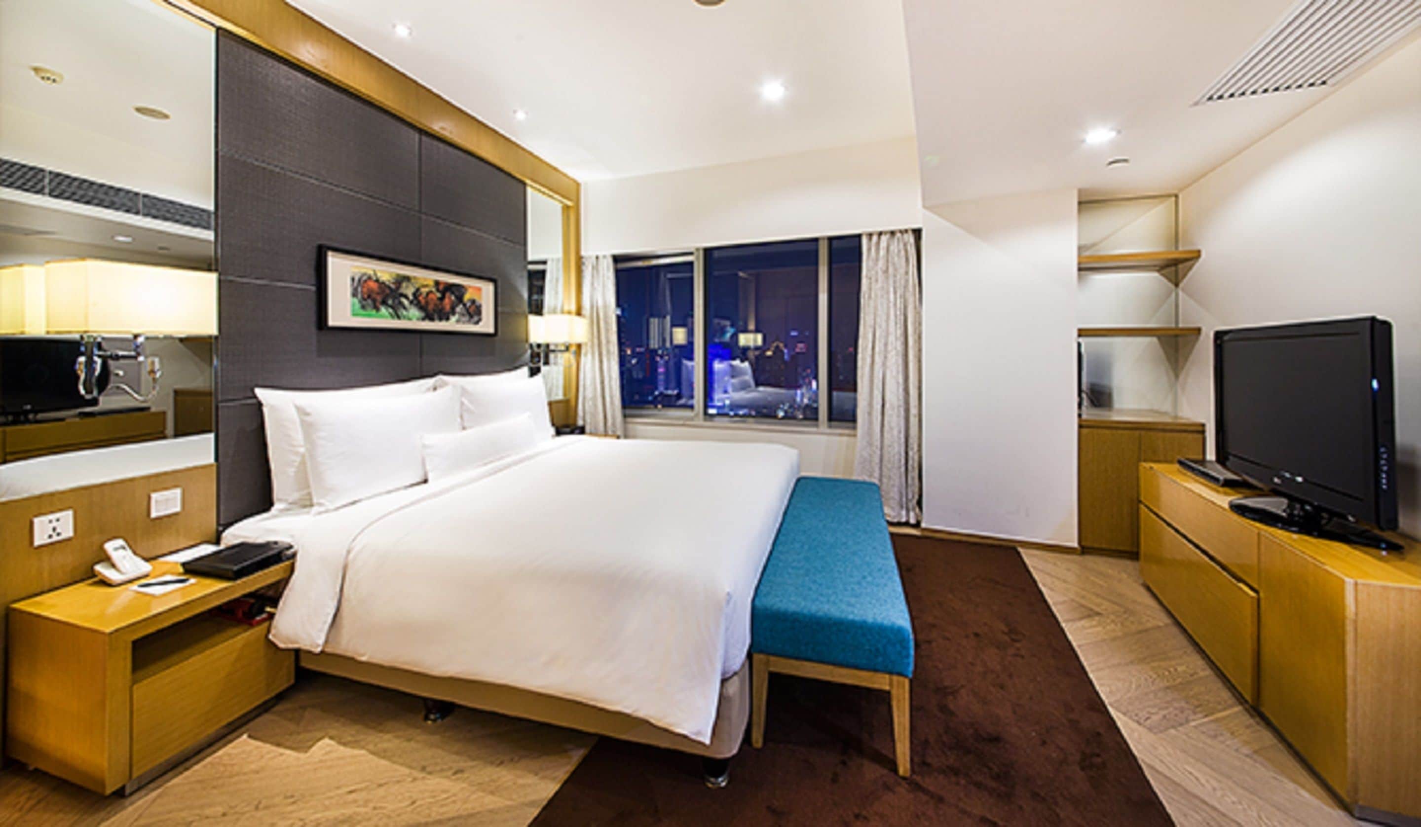 A hotel room with a large bed, a bedside table with a phone, a TV, and a window with a city view.