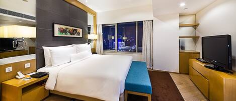A hotel room with a large bed, a bedside table with a phone, a TV, and a window with a city view.