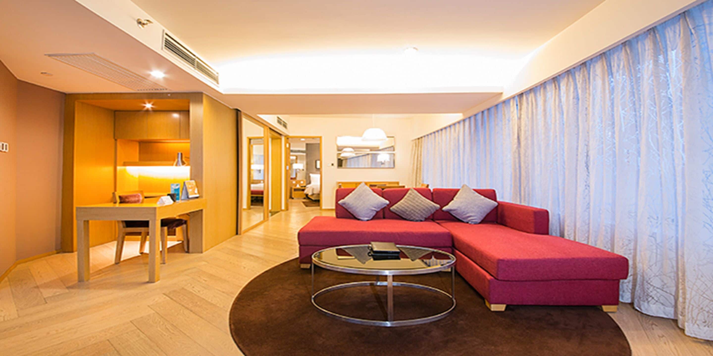 A modern hotel room with a red sofa, a round coffee table, and a dining area with a table and chairs.