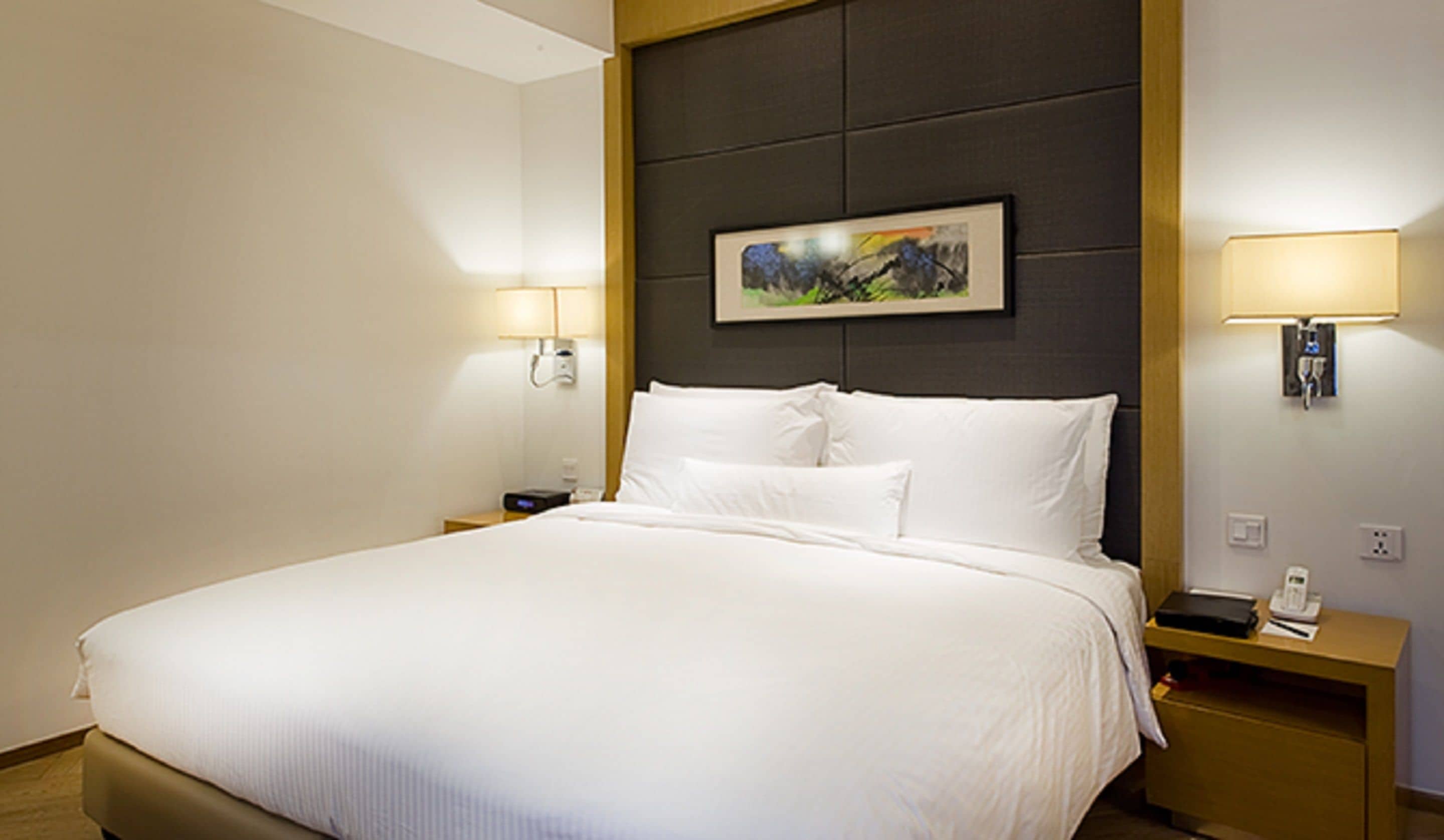 A hotel room with a large bed, two bedside tables, a painting above the headboard, and two wall-mounted lamps.