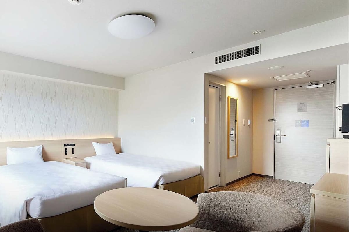 Habitación de hotel con dos camas, una mesa redonda, un sofá, un espejo y una puerta que da a un baño.