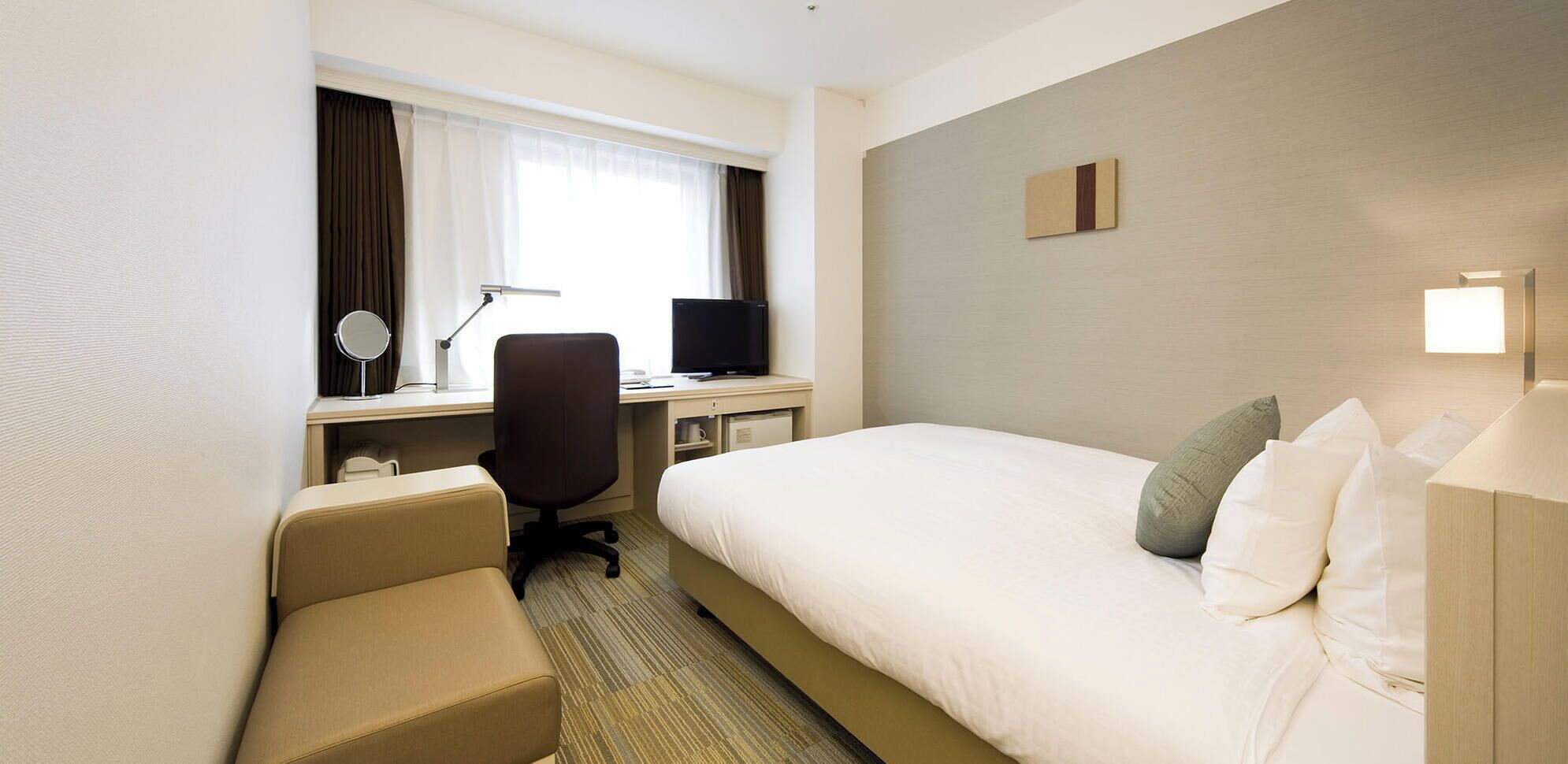 A hotel room with a bed, a desk with a computer, a chair, a TV, and a bedside table with a lamp.