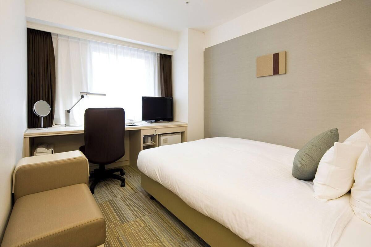 A hotel room with a bed, a desk with a computer, a chair, a TV, and a bedside table with a lamp.