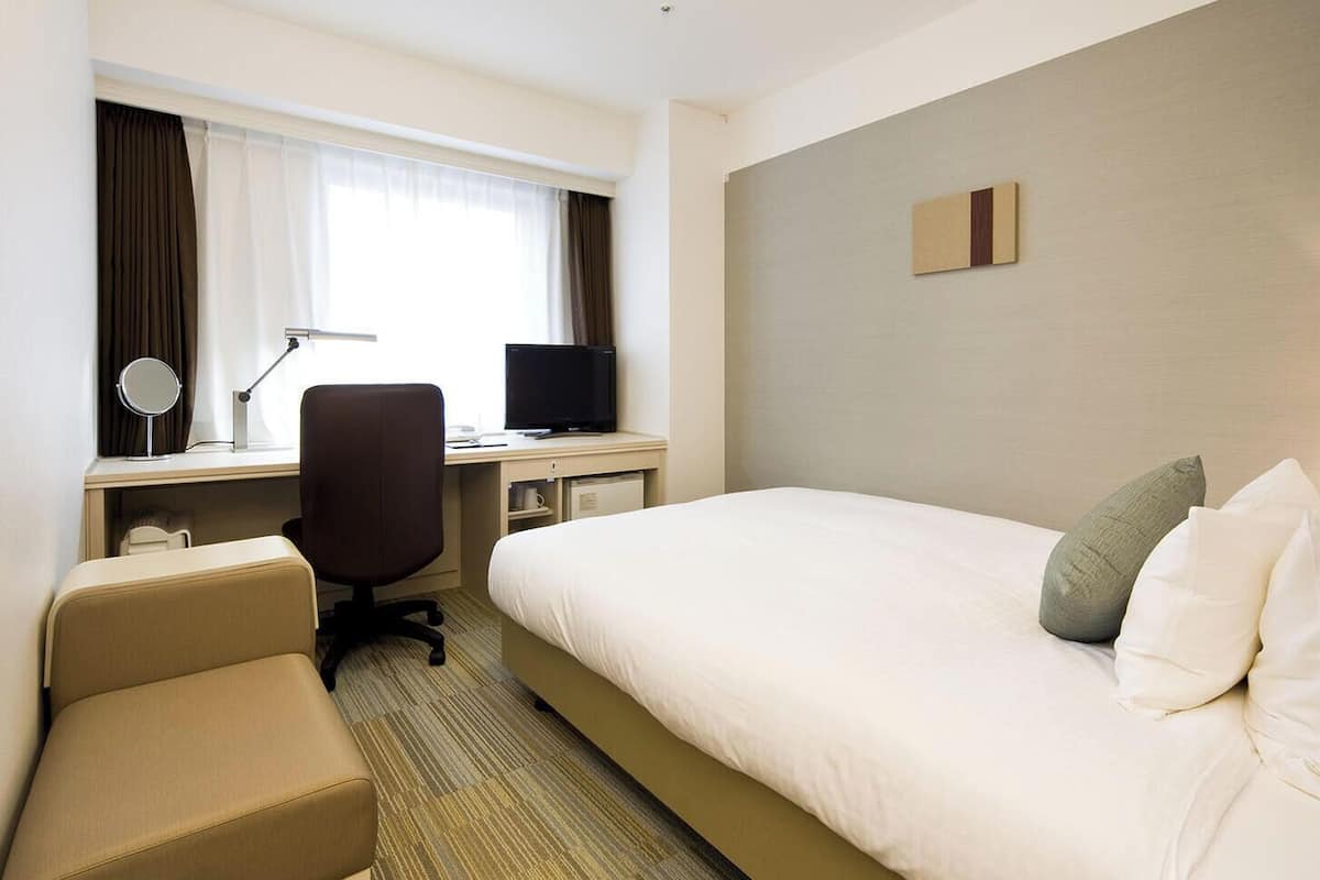 Habitación de hotel con cama, escritorio con computadora, silla, televisor y mesita de noche con lámpara.