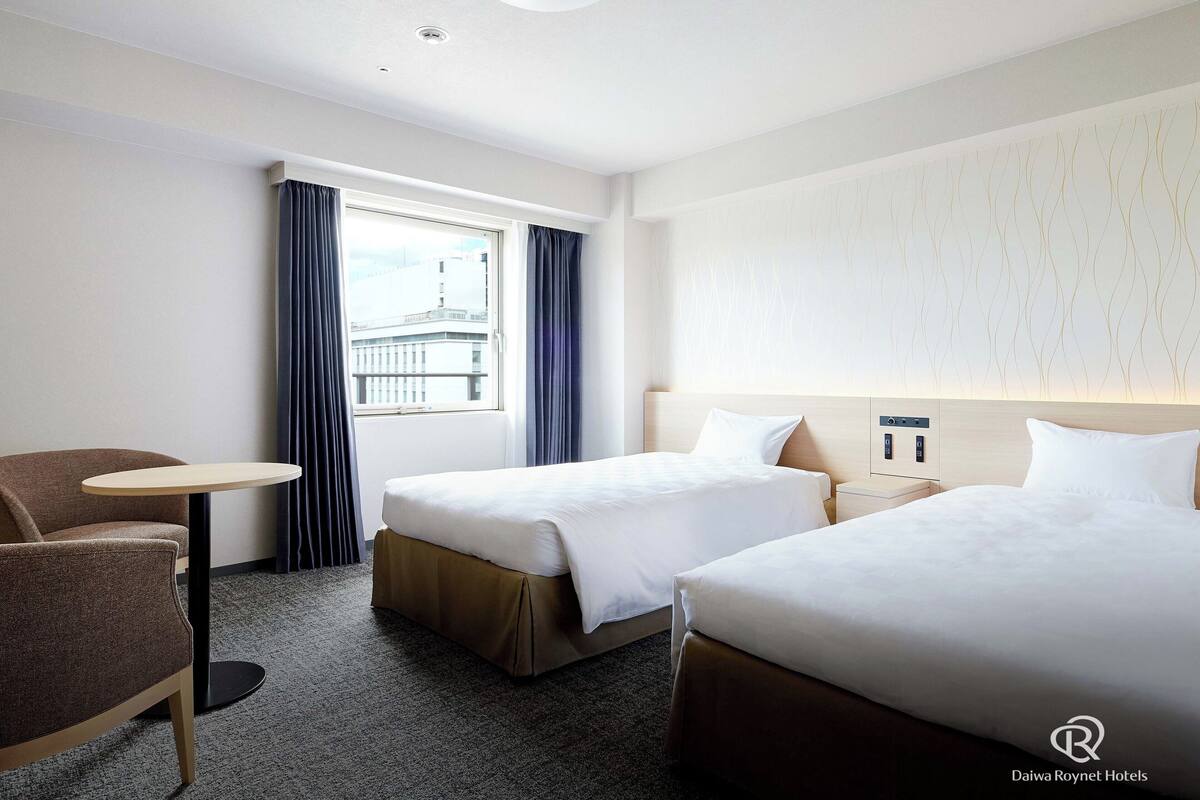A hotel room with two beds, a small table, a chair, and a window with curtains.