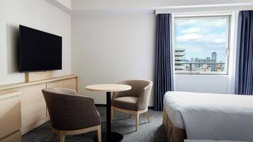 Una habitación de hotel con cama, escritorio, silla, una pequeña mesa redonda, televisor y vista a la ciudad desde la ventana.
