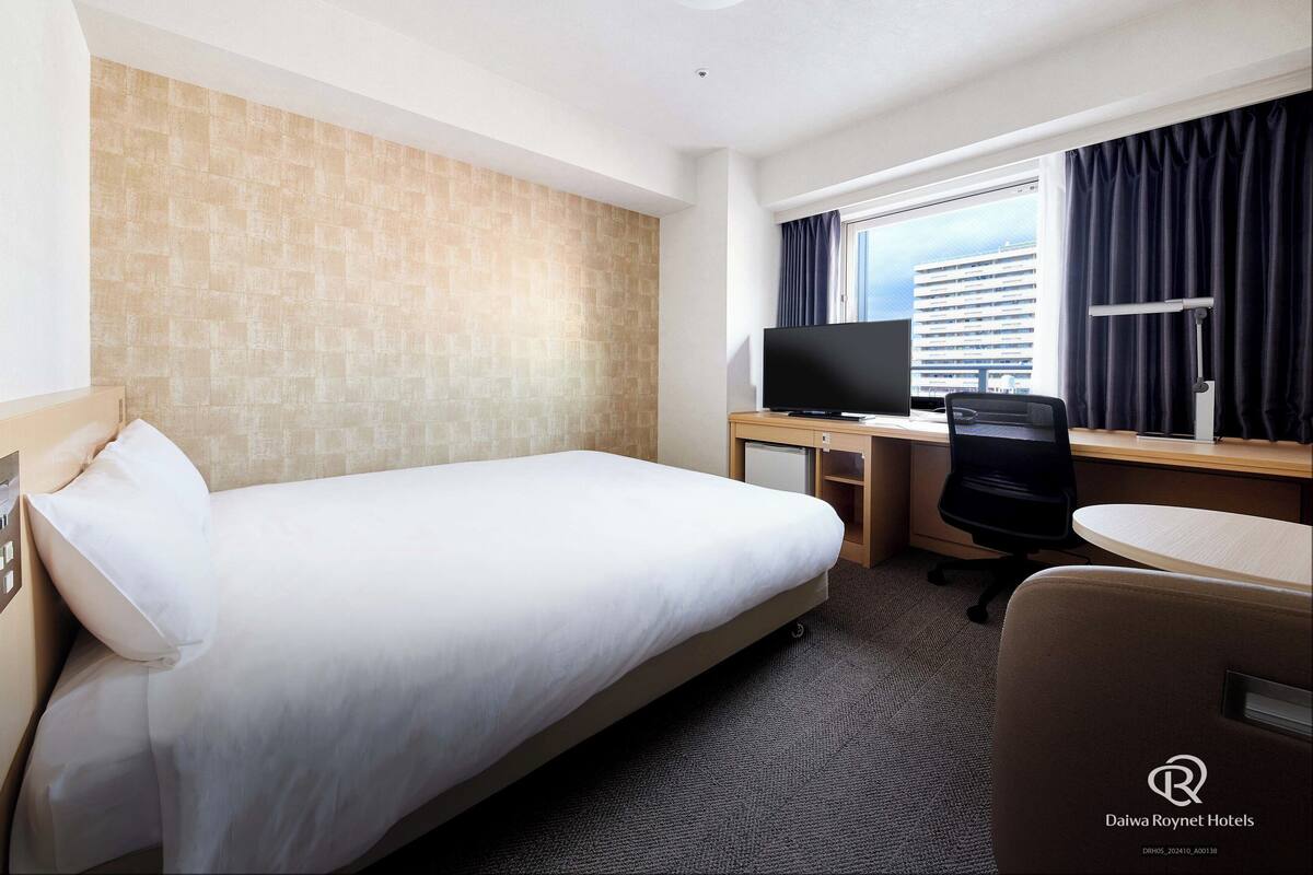 A hotel room with a bed, a desk with a computer, a chair, a TV, and a window with curtains.