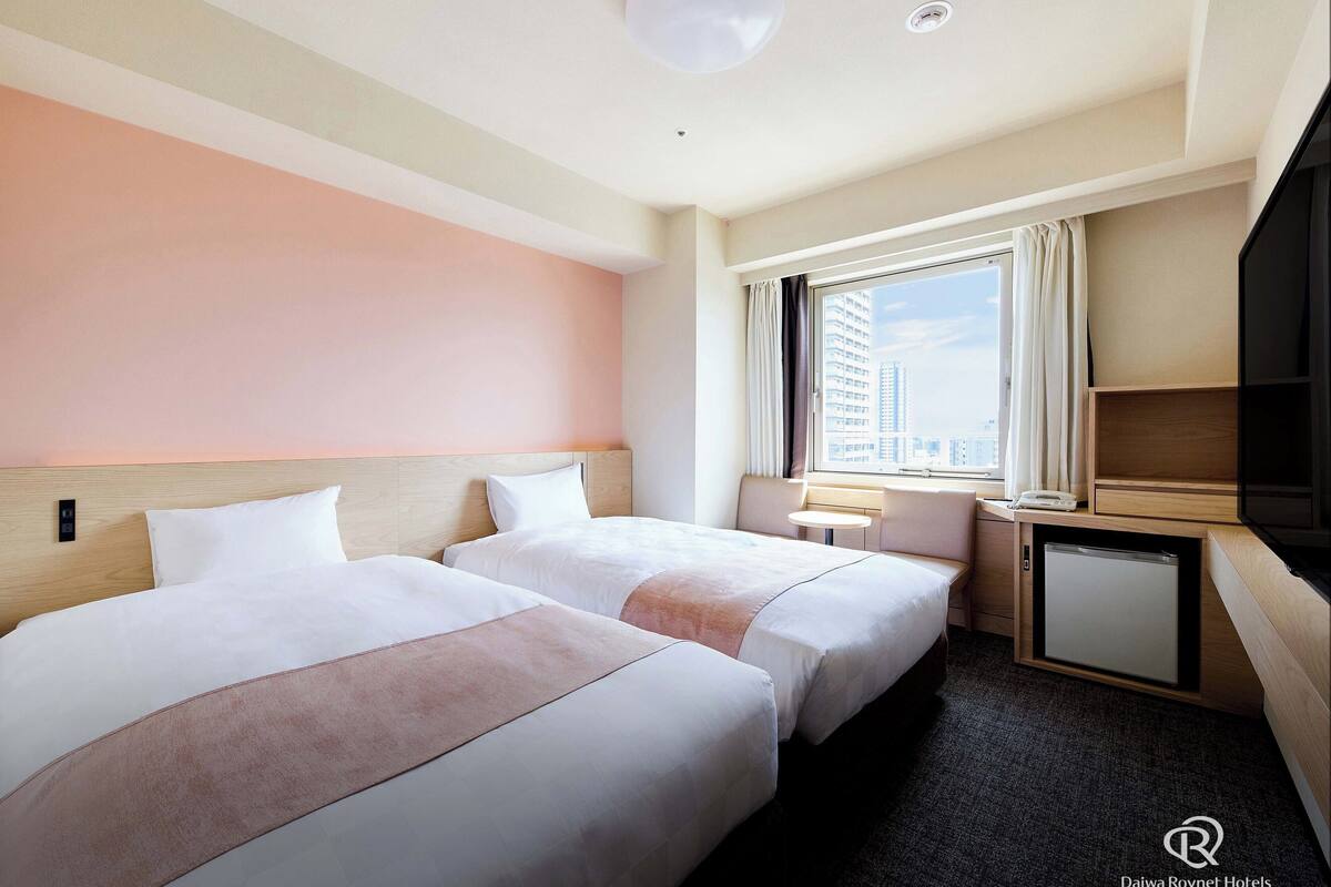 A hotel room with two beds, a small table, a TV, and a window with a city view.