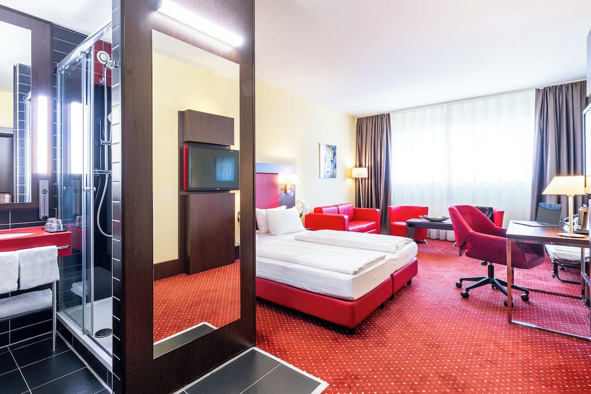 Une chambre d’hôtel avec un tapis rouge, un lit, un bureau avec une chaise, une télévision et un espace douche.