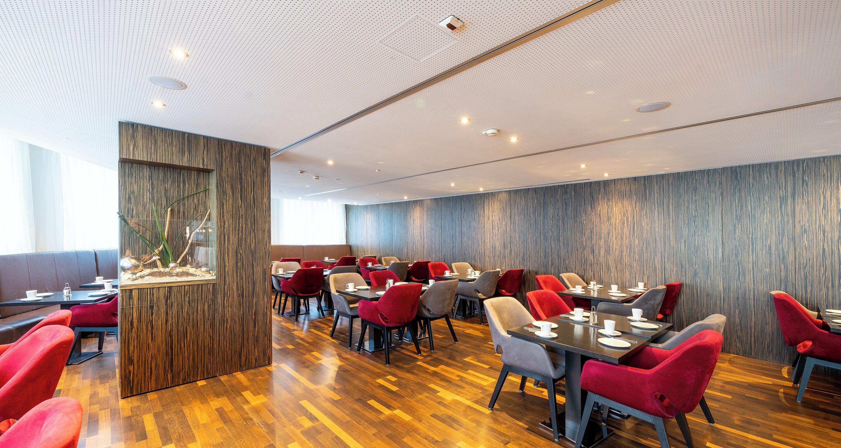 Un restaurante moderno con suelo de madera, sillas rojas y grises, y una planta decorativa en el centro.