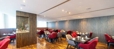 Un restaurante moderno con suelo de madera, sillas rojas y grises, y una planta decorativa en el centro.