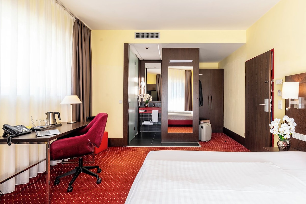Habitación de hotel con cama, escritorio con teléfono, una silla, una mesita con lámpara y baño visible a través de una puerta abierta.