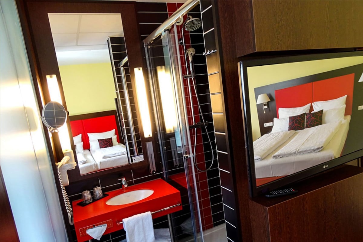 Habitación de hotel con una decoración en rojo y blanco, un espejo grande, un lavamanos rojo, un televisor y una zona de ducha.