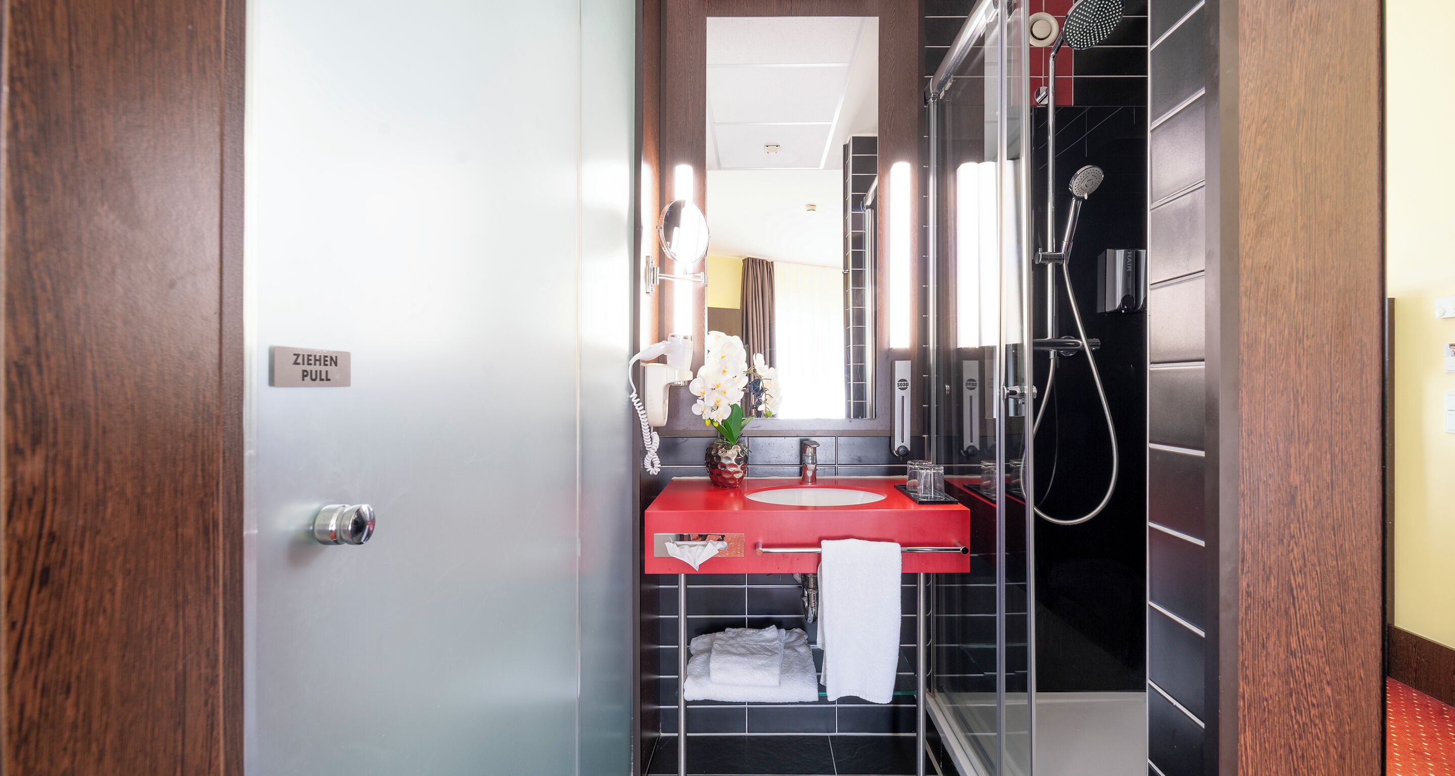 Une salle de bain moderne avec un lavabo rouge, une douche en verre et un miroir à reflet circulaire.