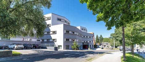 Ein modernes Gebäude mit flachem Dach und großen Fenstern, umgeben von Bäumen und parkenden Autos.