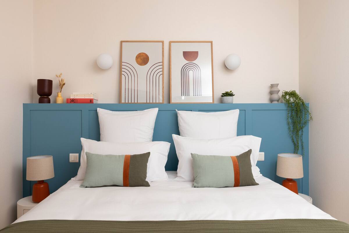 A neatly made bed with white and green pillows, a blue headboard, and two framed artworks above.