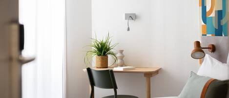 A modern hotel room with a bed, a desk with a plant, a chair, and a wall-mounted light fixture.