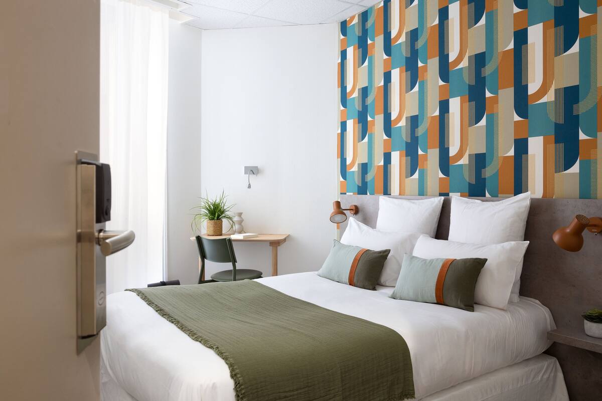 A hotel room with a bed, a desk, a chair, a potted plant, and a wall with a geometric pattern.
