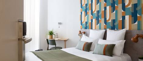 A hotel room with a bed, a desk, a chair, a potted plant, and a wall with a geometric pattern.
