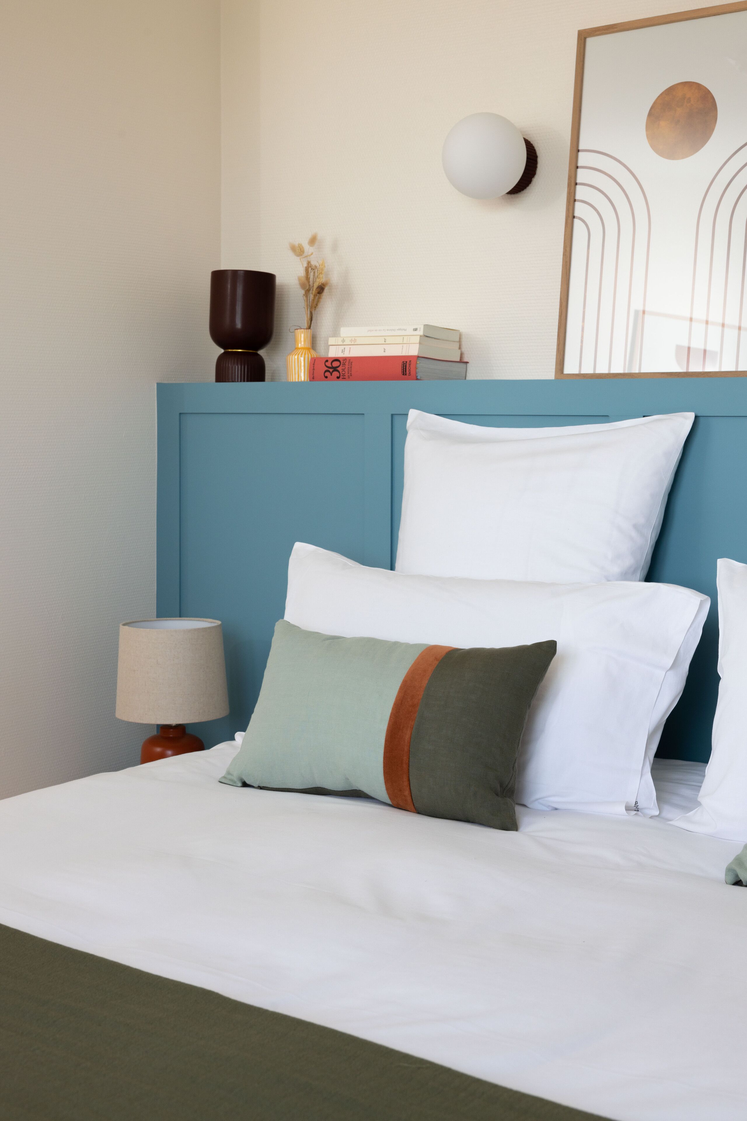 A neatly made bed with white bedding and multiple pillows, a bedside lamp, and a wall-mounted light fixture.