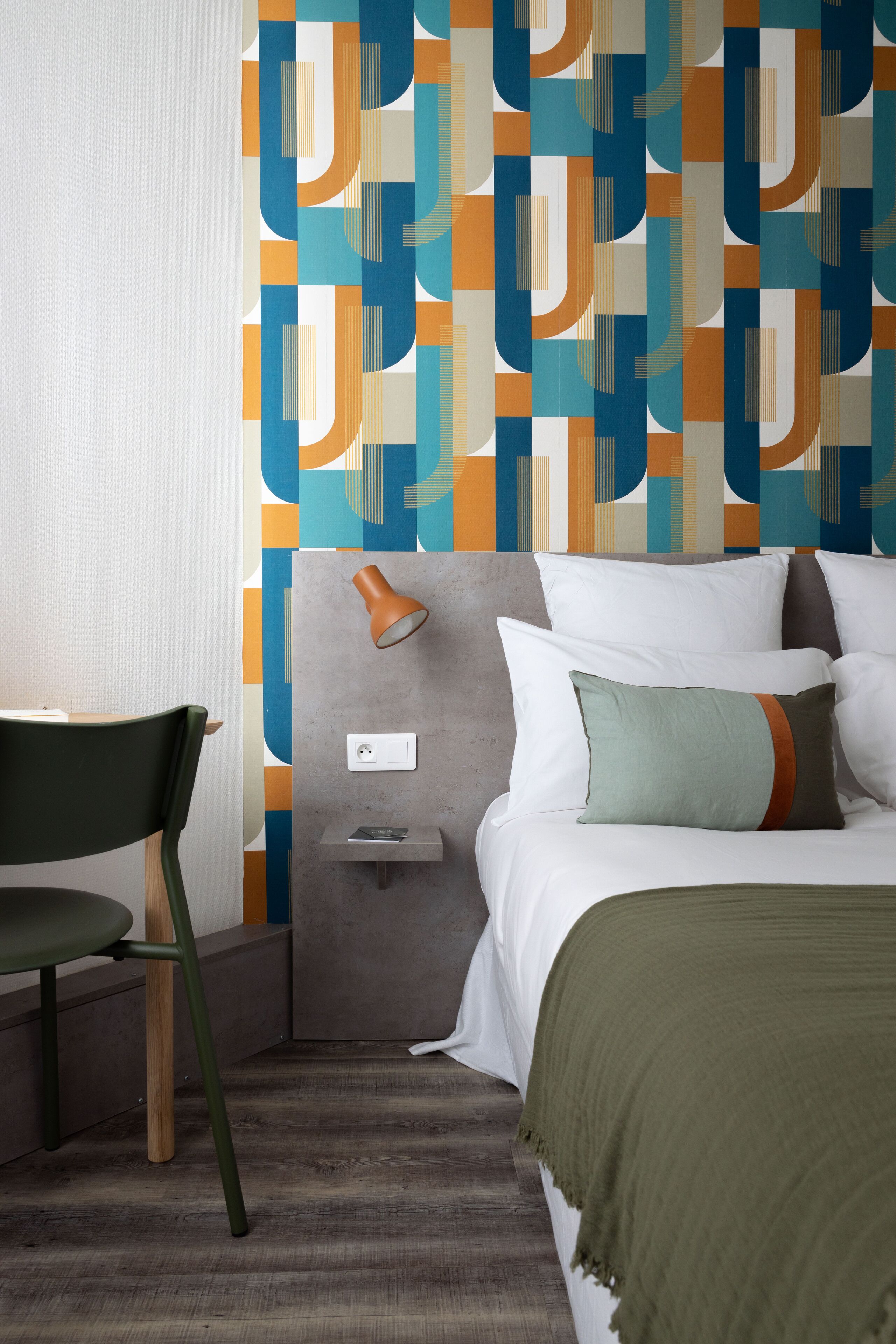 Ein Hotelzimmer mit modernem Design, ausgestattet mit einem Bett mit weißen Leinentüchern, einem grünen Sessel, einem Nachttisch mit Lampe und einer Wand mit geometrischem Muster.