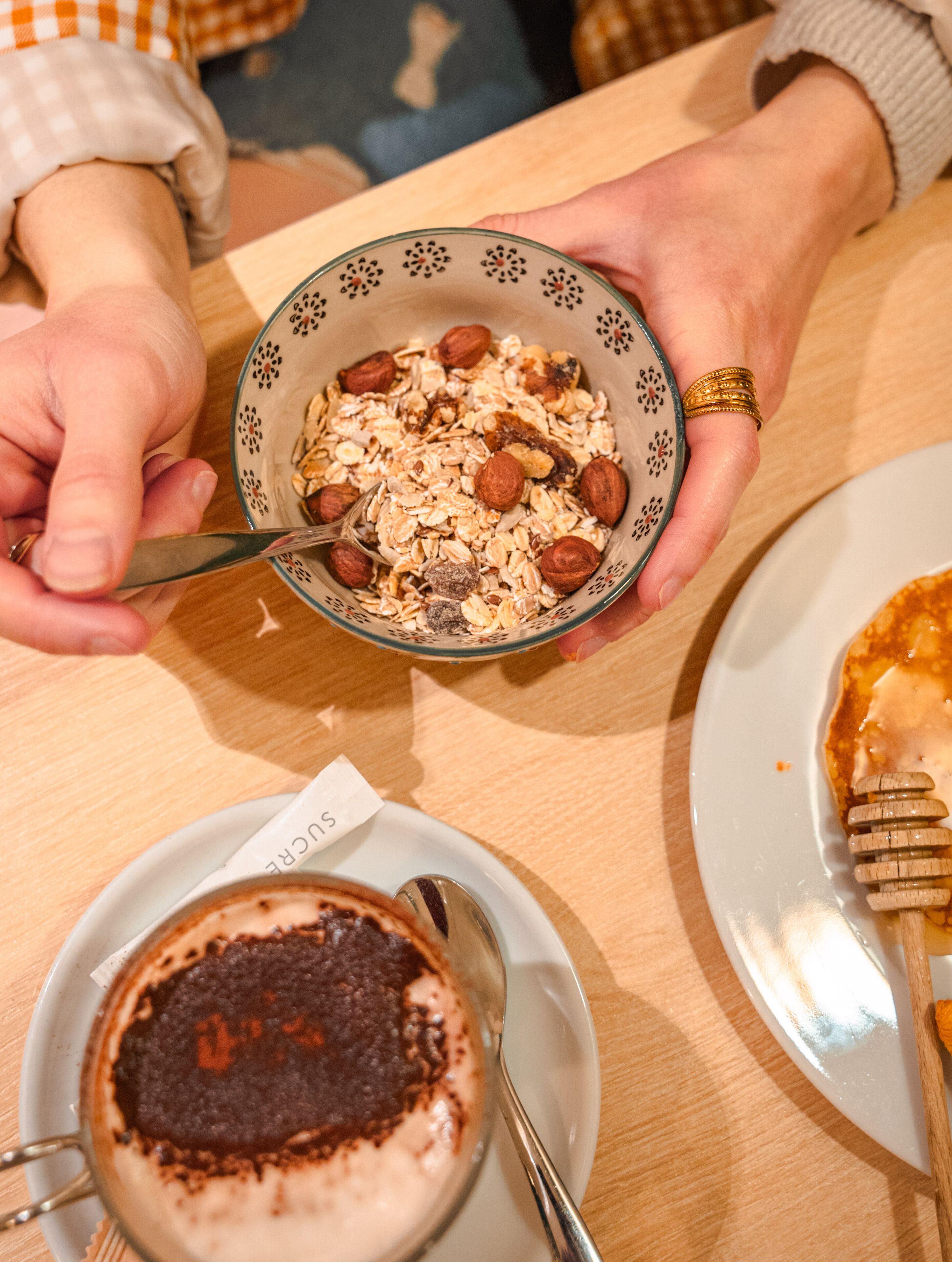 Un tazón de cereal con nueces y una cuchara, un plato con un postre de chocolate y un recipiente para miel sobre una mesa.