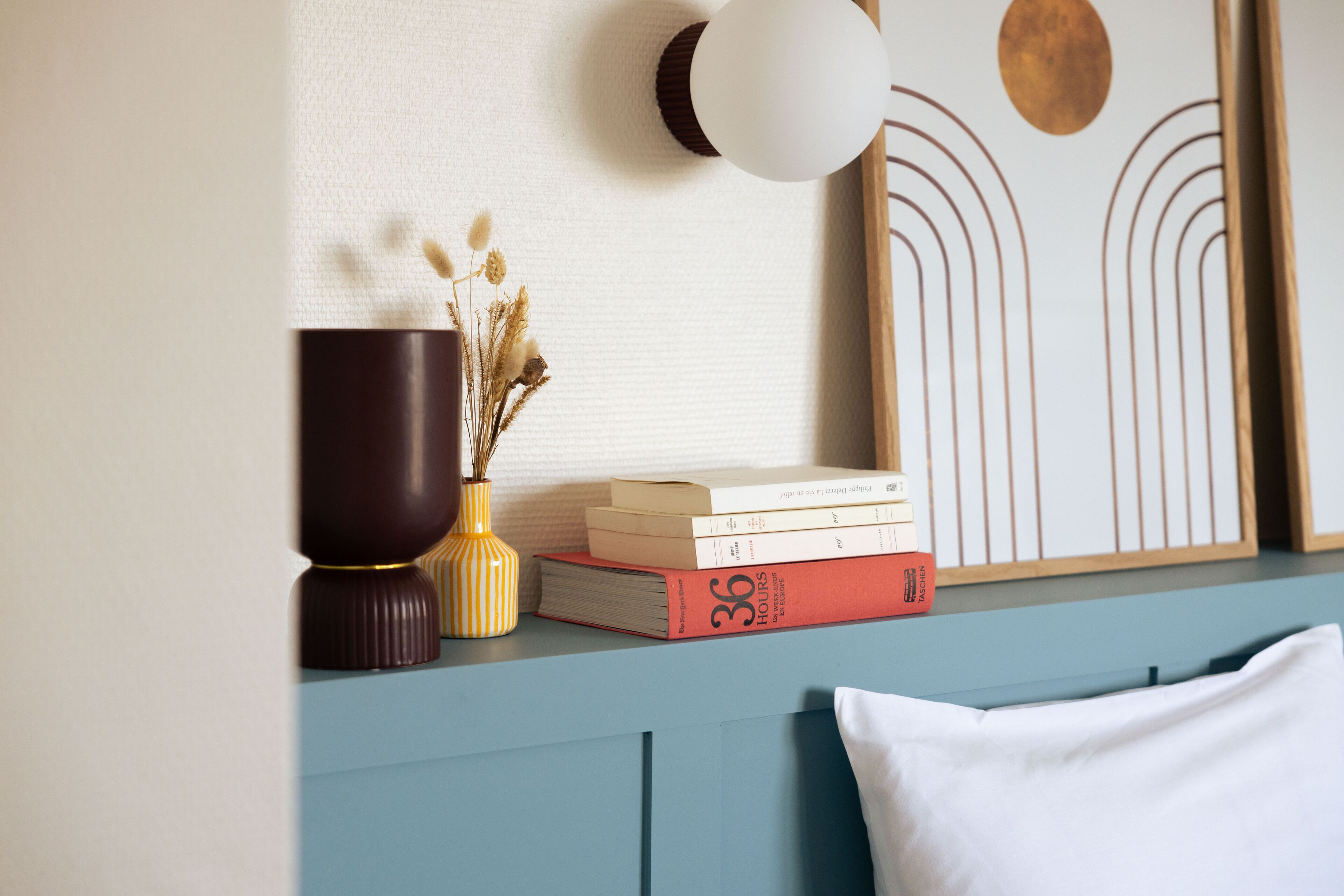 A neatly arranged shelf with books, a vase with dried flowers, and a wall-mounted light fixture.