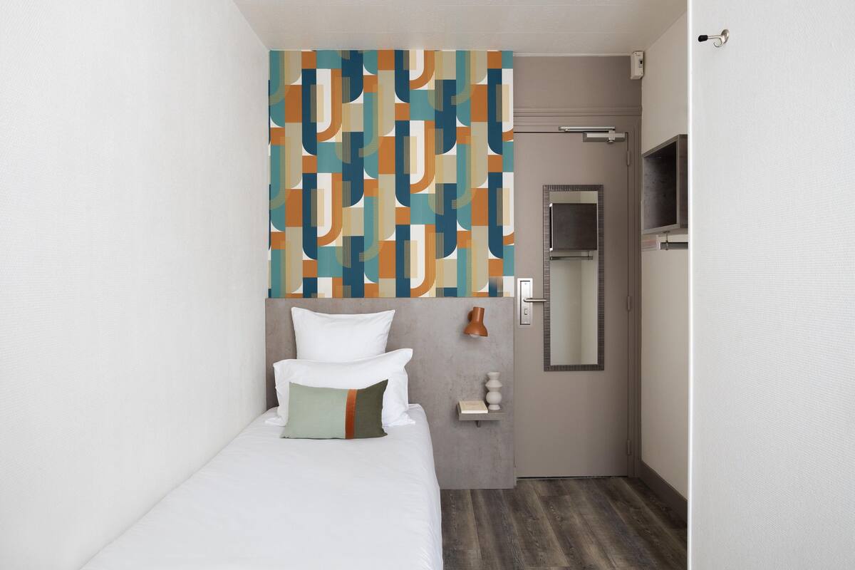 A small, modern hotel room with a bed, a mirror, a TV, and a patterned curtain.