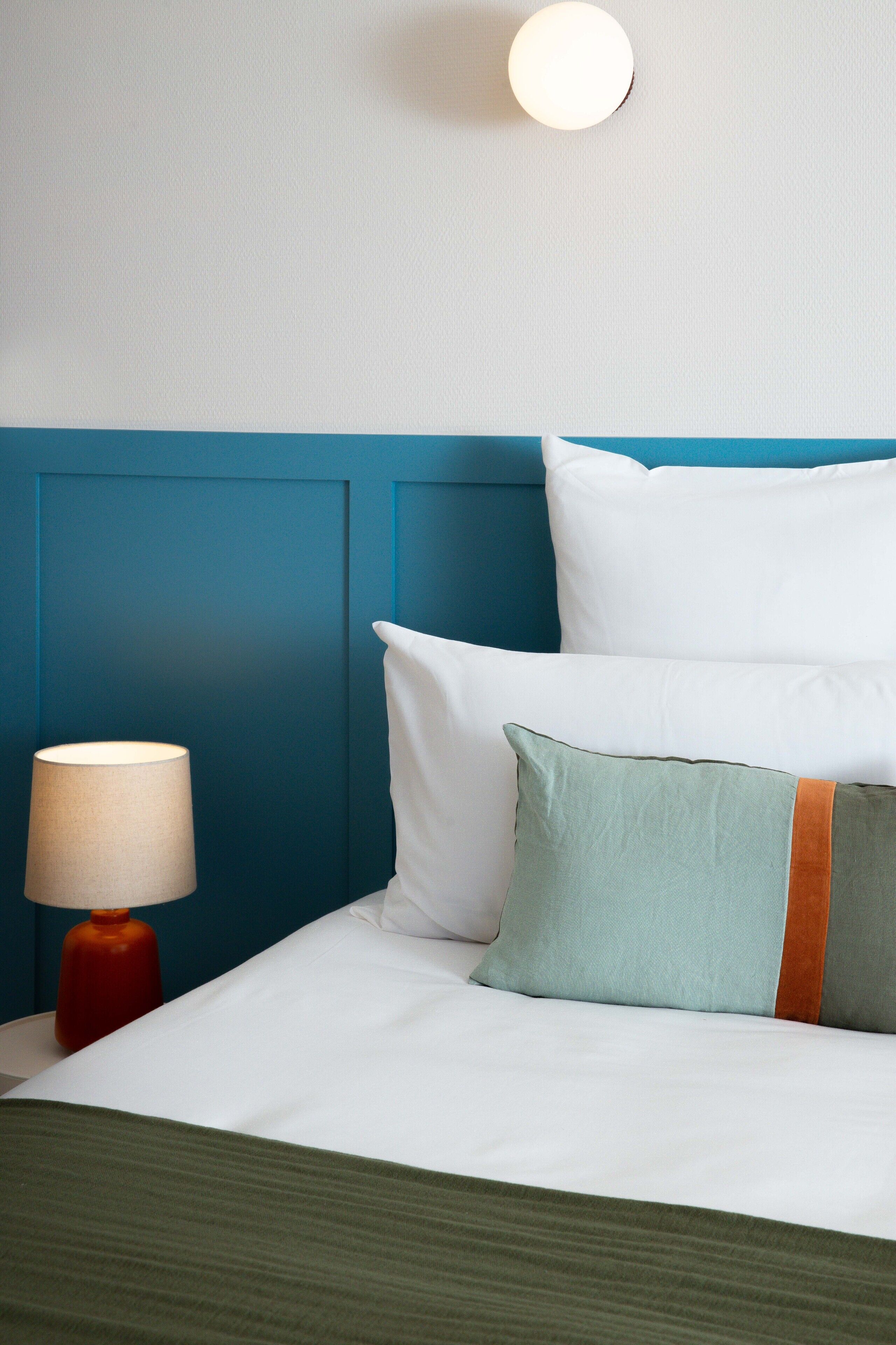 A neatly made bed with white and green pillows, a teal headboard, and a red lamp on a white nightstand.