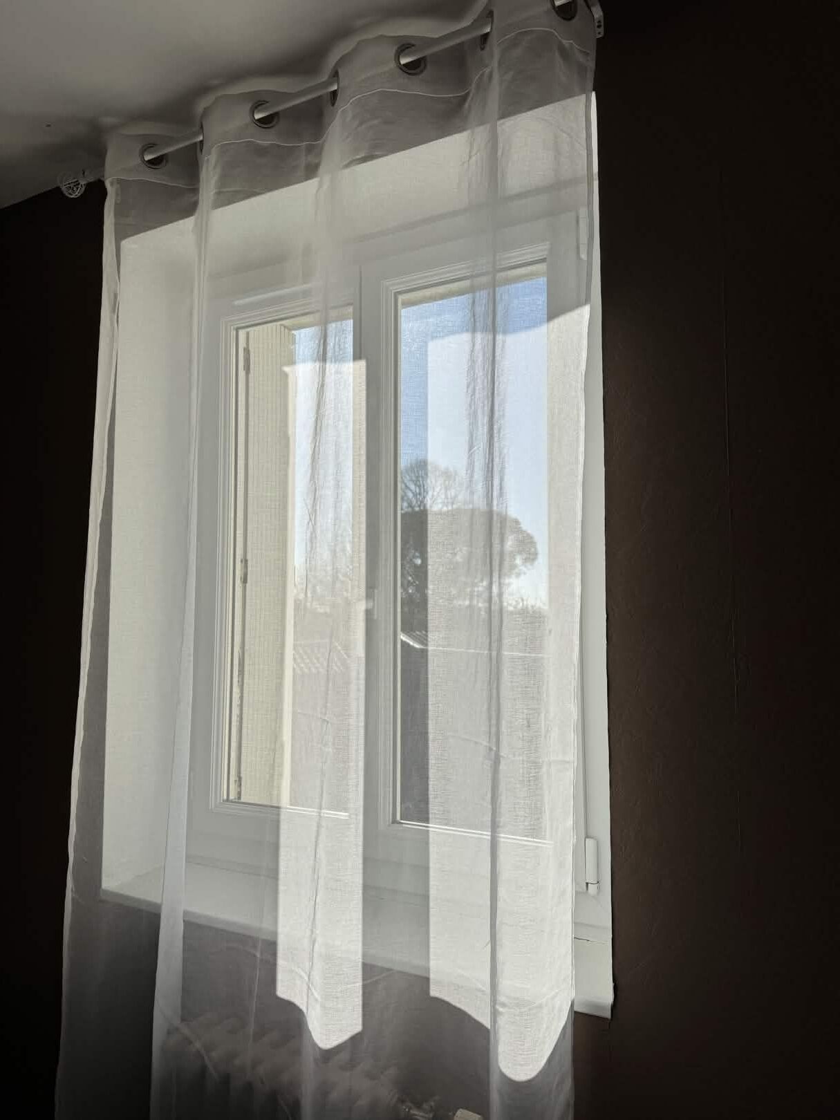 Janela com cortina translúcida, moldura branca e vista para uma árvore lá fora.