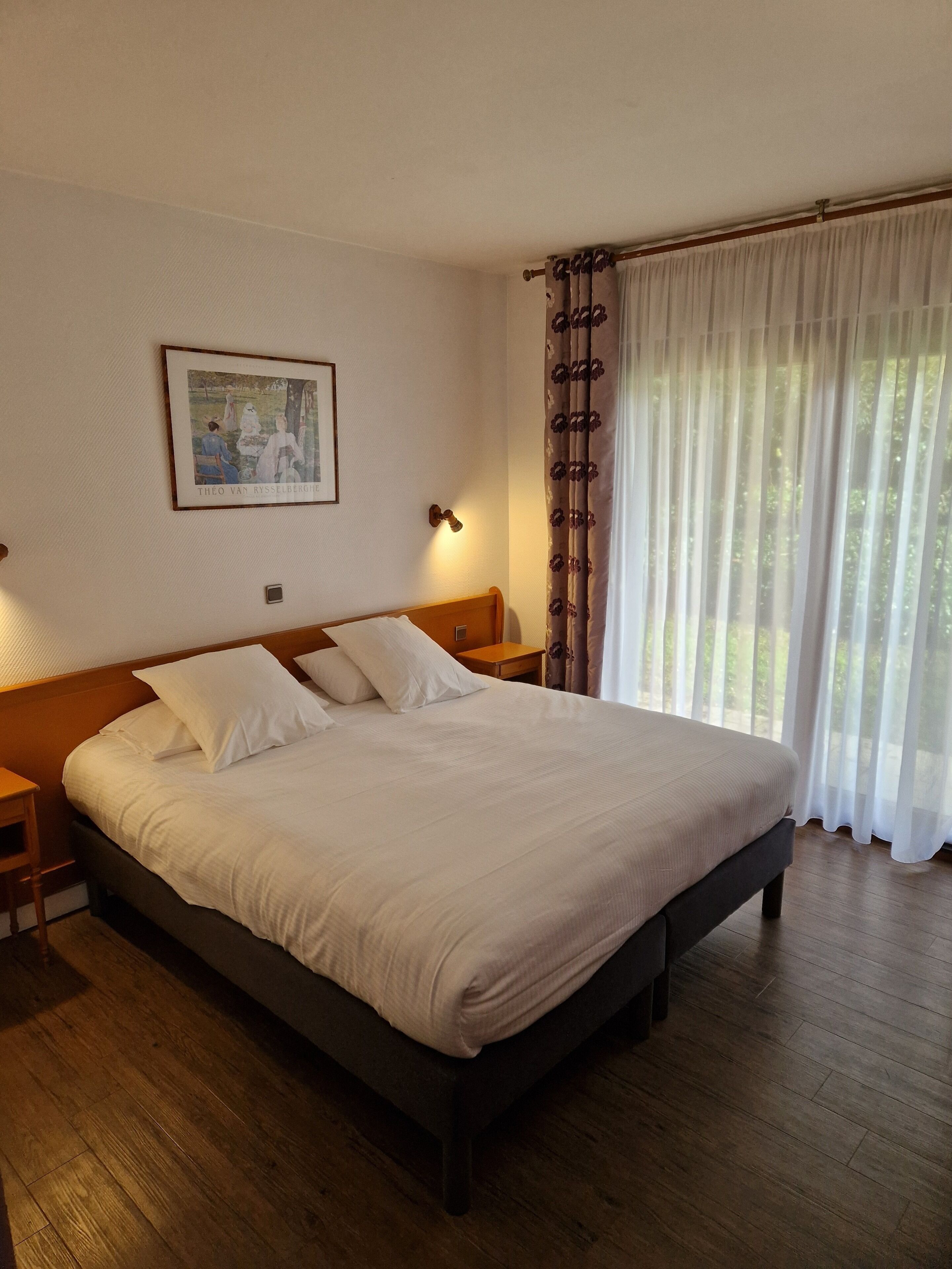 Una cama bien tendida en una habitación de hotel, con un cabecero de madera, un cuadro enmarcado en la pared y una ventana con cortinas de gasa.