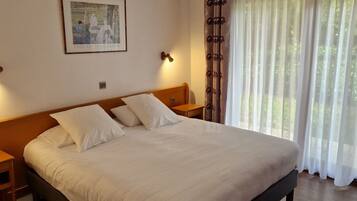 Una cama bien tendida en una habitación de hotel, con un cabecero de madera, un cuadro enmarcado en la pared y una ventana con cortinas de gasa.
