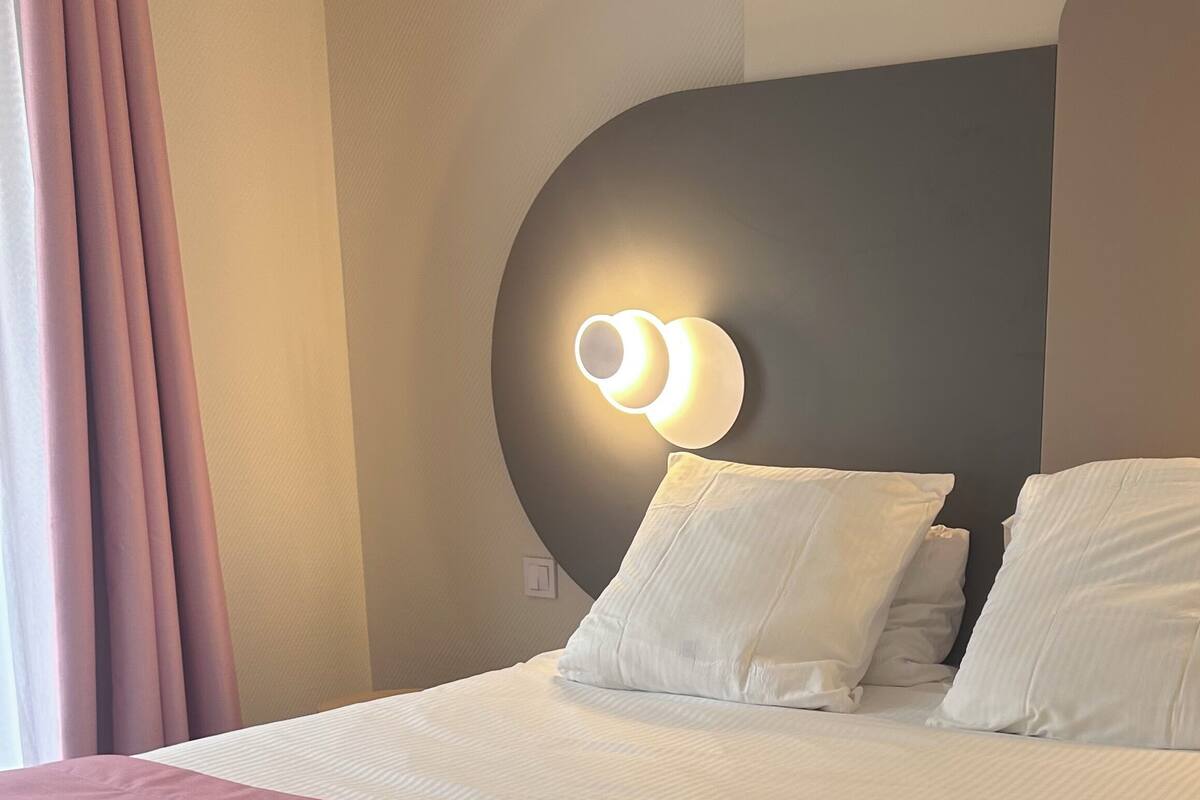 A neatly made bed with white and pink bedding, a round wall-mounted light fixture, and a window with curtains.