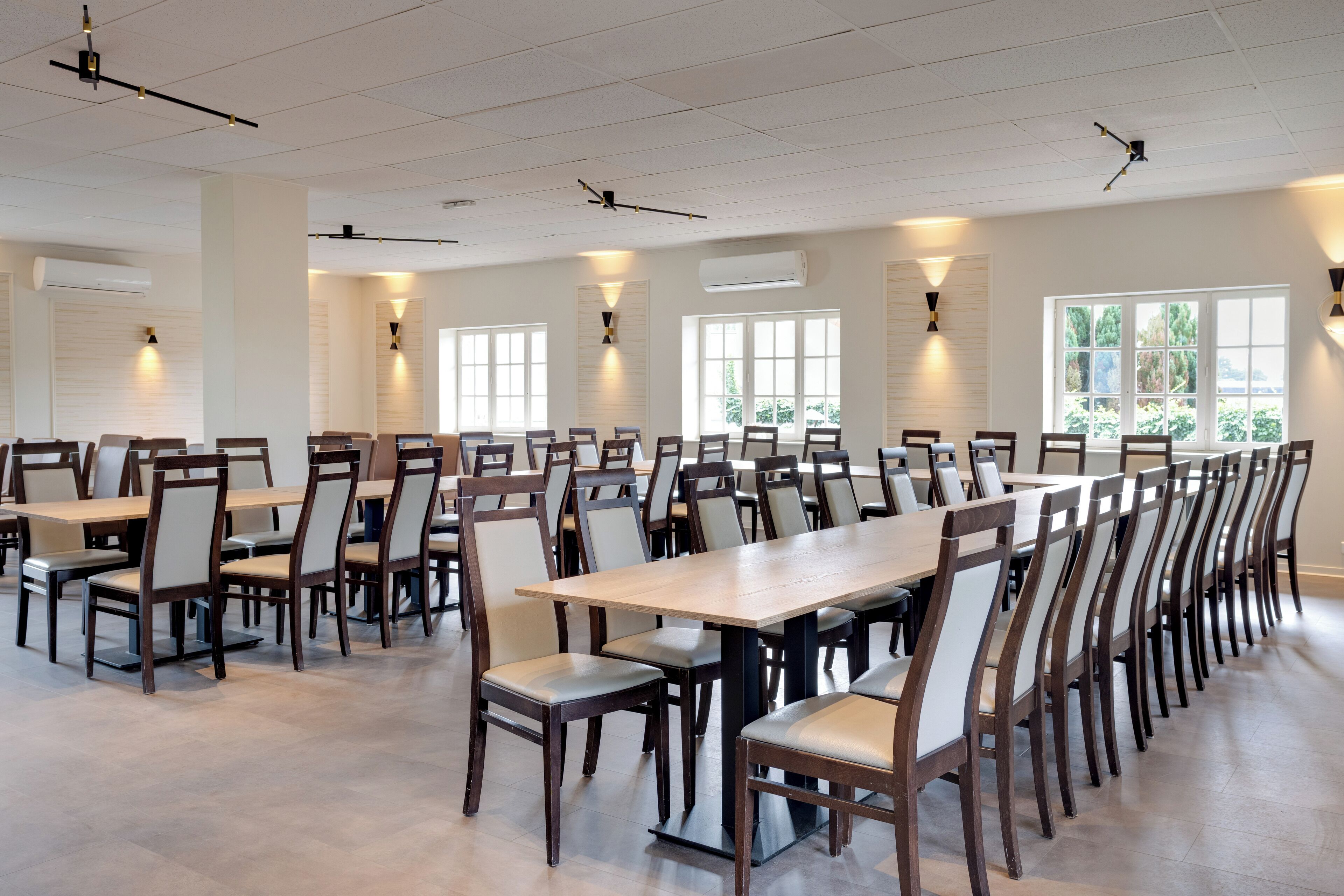 Un comedor espacioso con largas mesas y sillas de madera, ventanas amplias que dejan entrar luz natural, y lámparas de techo modernas.