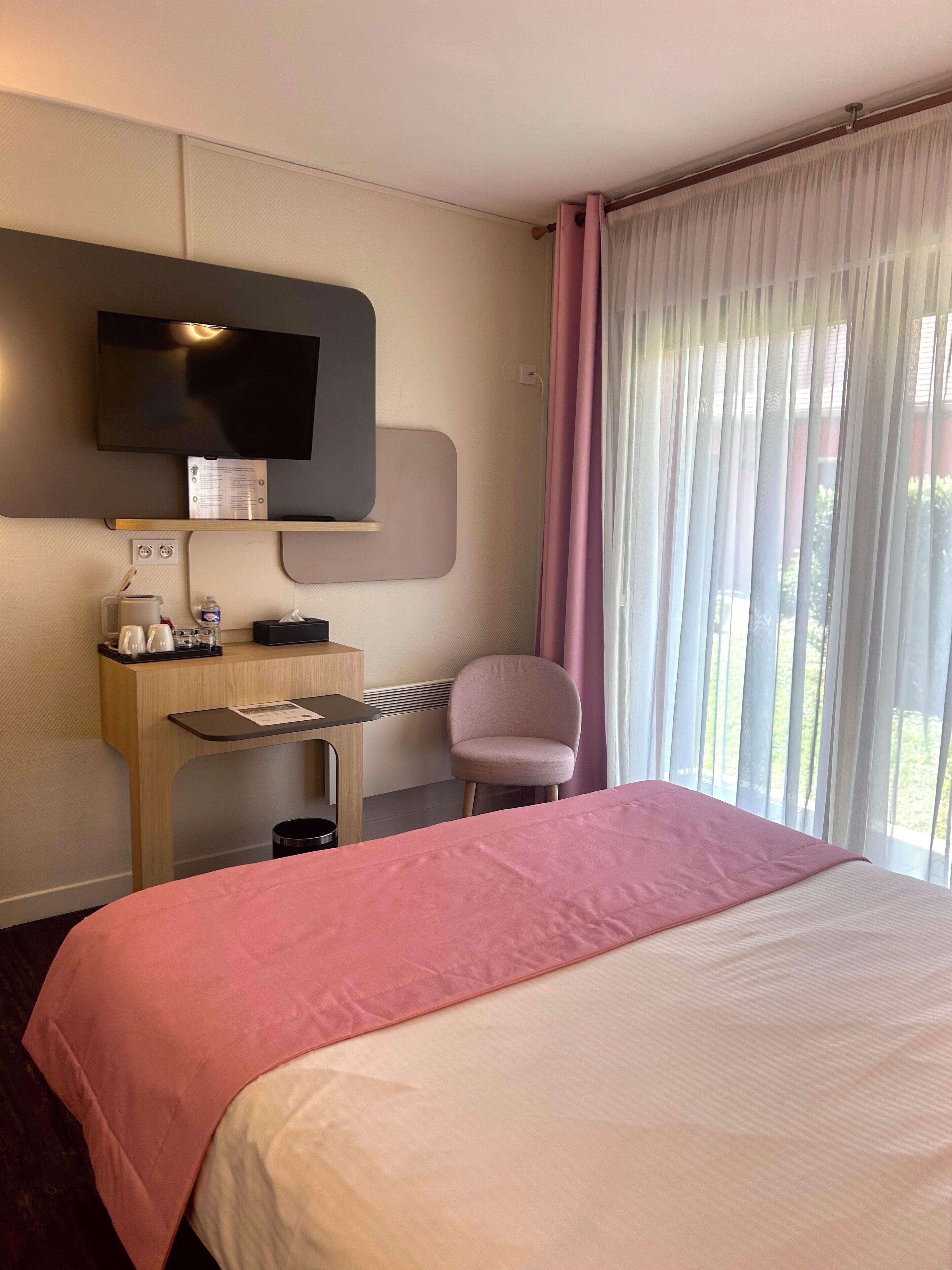 Habitación de hotel con cama, escritorio, silla, televisor montado en la pared y ventana con cortinas.